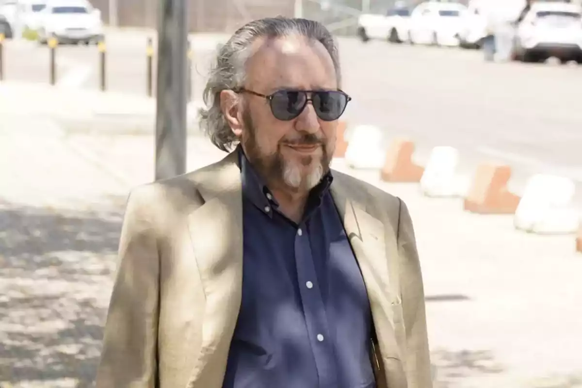 Hombre de cabello canoso y barba con gafas de sol, vistiendo saco beige y camisa azul, caminando al aire libre en una zona urbana