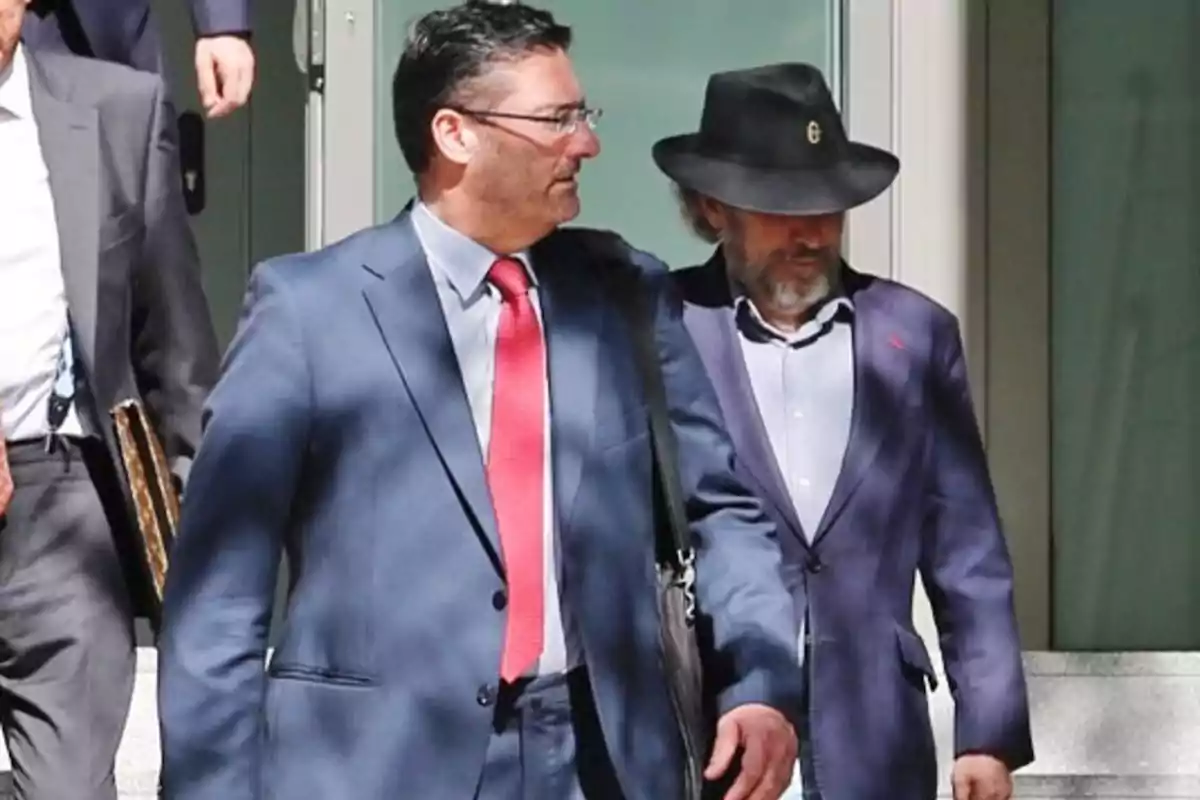 Dos hombres vestidos con traje caminan juntos por la calle uno de ellos lleva sombrero negro y el otro una corbata roja Dos hombres vestidos con traje caminan juntos por la calle uno de ellos lleva sombrero negro y el otro una corbata roja
