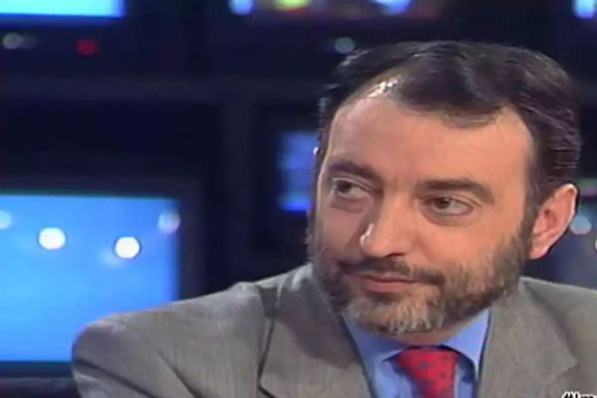 Hombre de barba con traje gris y corbata roja en un estudio de televisión con pantallas al fondo Hombre de barba con traje gris y corbata roja en un estudio de televisión con pantallas al fondo