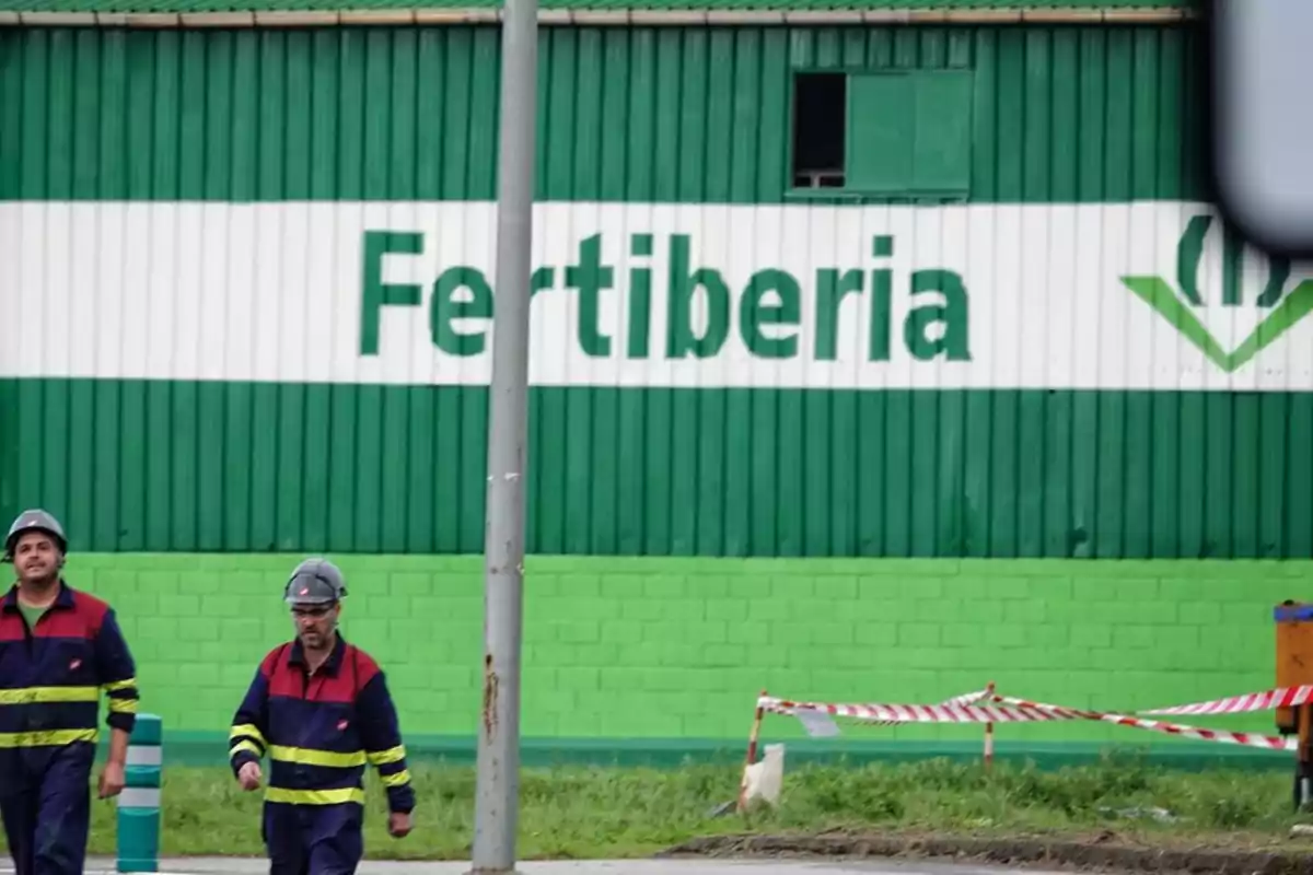 Dos trabajadores con casco y uniforme de seguridad caminan frente a un edificio verde con el letrero Fertiberia