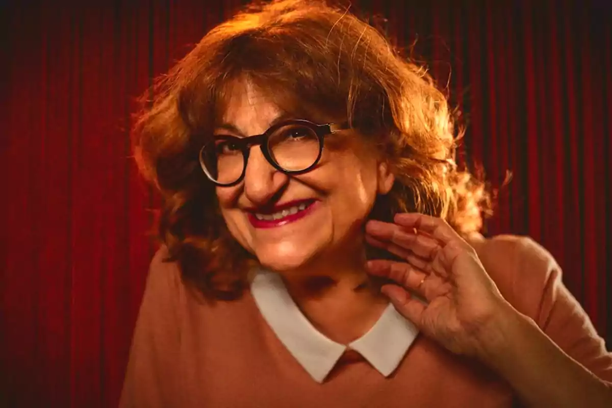 Mujer de cabello rizado y gafas grandes sonriendo frente a una cortina roja