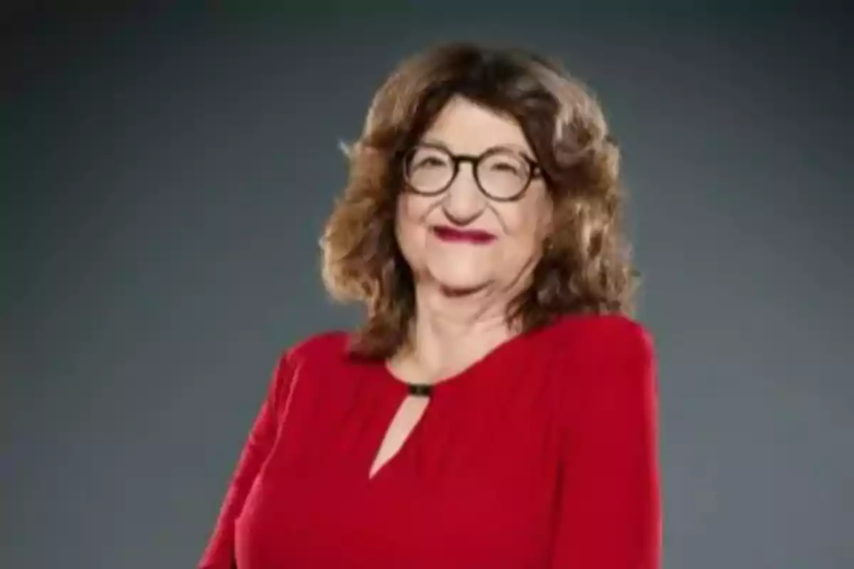 Mujer de cabello castaño y rizado con gafas y vestido rojo posando frente a un fondo gris Mujer de cabello castaño y rizado con gafas y vestido rojo posando frente a un fondo gris