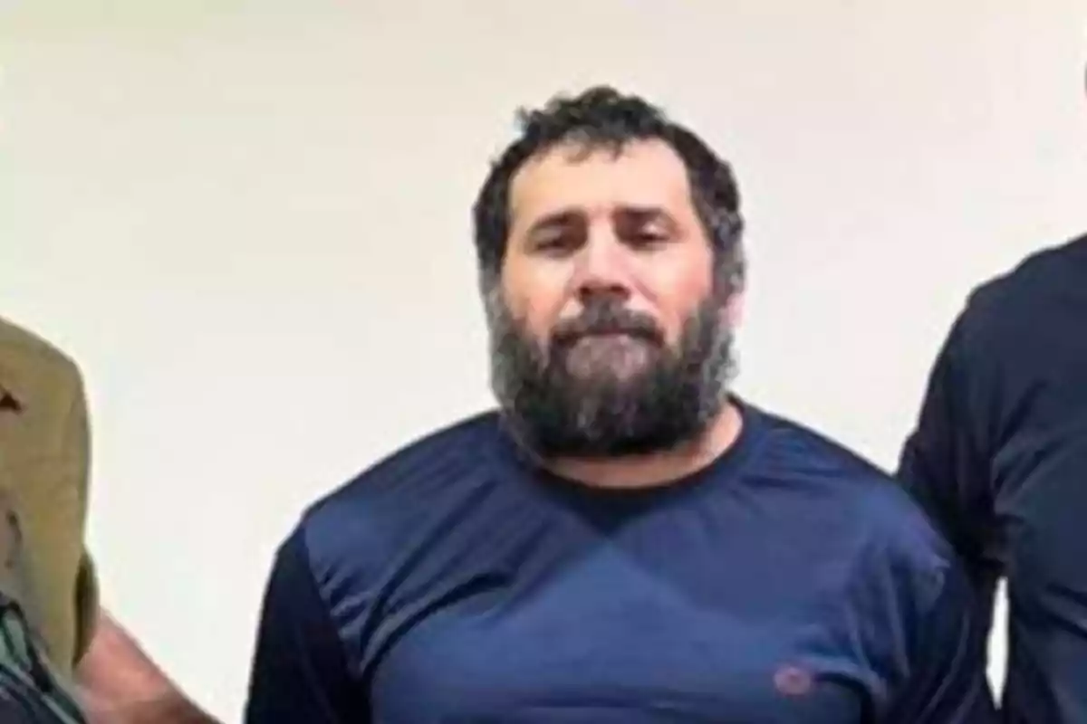 Hombre de barba y cabello oscuro con camiseta azul de manga larga de pie frente a una pared clara