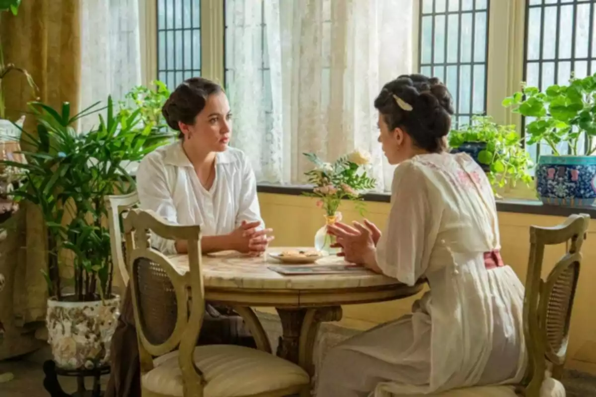 Dos mujeres vestidas con ropa de época conversan sentadas en una mesa redonda junto a una ventana rodeadas de plantas y luz natural