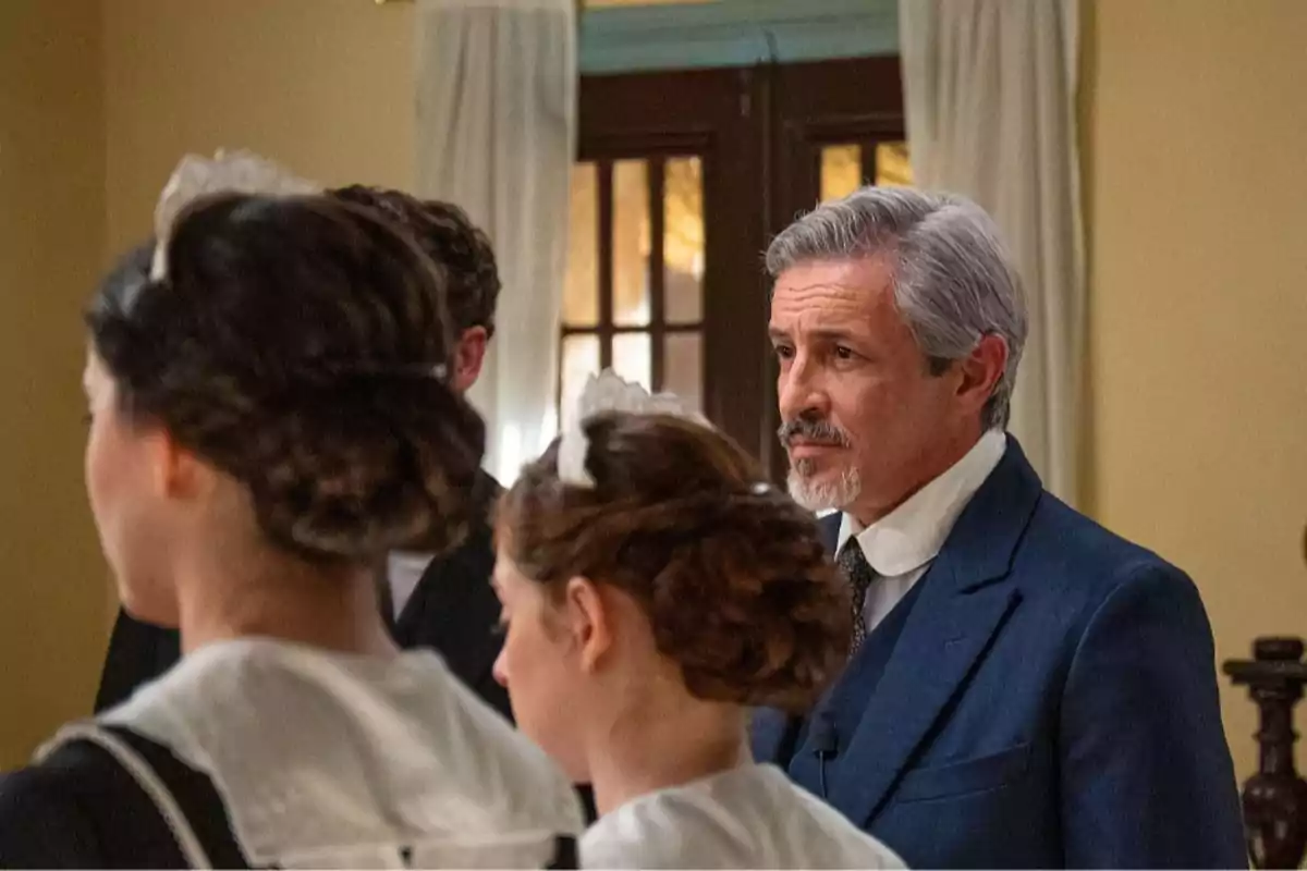 Cristóbal (Fernando Coronado) de cabello canoso y barba corta viste un traje azul mientras observa a tres personas con uniformes de sirvienta en una habitación iluminada con cortinas blancas. Cristóbal (Fernando Coronado) de cabello canoso y barba corta viste un traje azul mientras observa a tres personas con uniformes de sirvienta en una habitación iluminada con cortinas blancas.