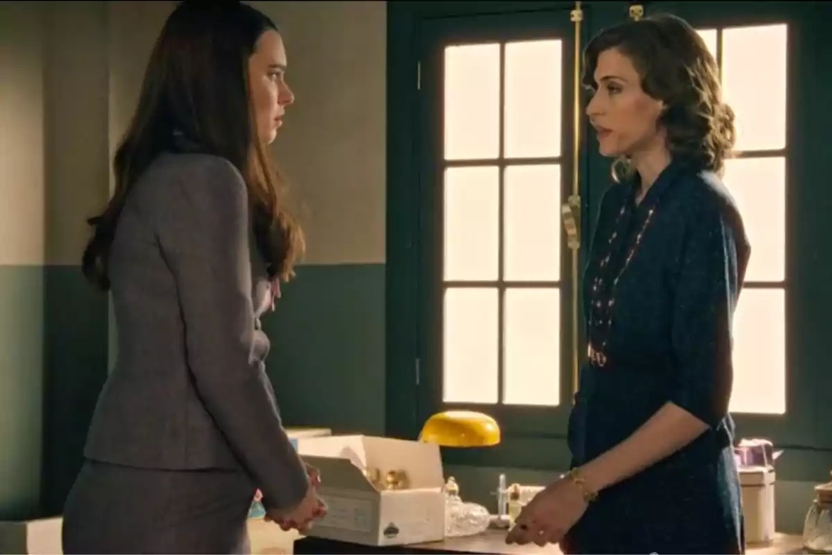Marta de la Reina (Marta Belmonte) y Fina Valero (Alba Brunet) conversan de pie en una oficina iluminada por la luz que entra por una ventana, una de ellas lleva un traje gris y la otra un vestido azul oscuro. Marta de la Reina (Marta Belmonte) y Fina Valero (Alba Brunet) conversan de pie en una oficina iluminada por la luz que entra por una ventana, una de ellas lleva un traje gris y la otra un vestido azul oscuro.