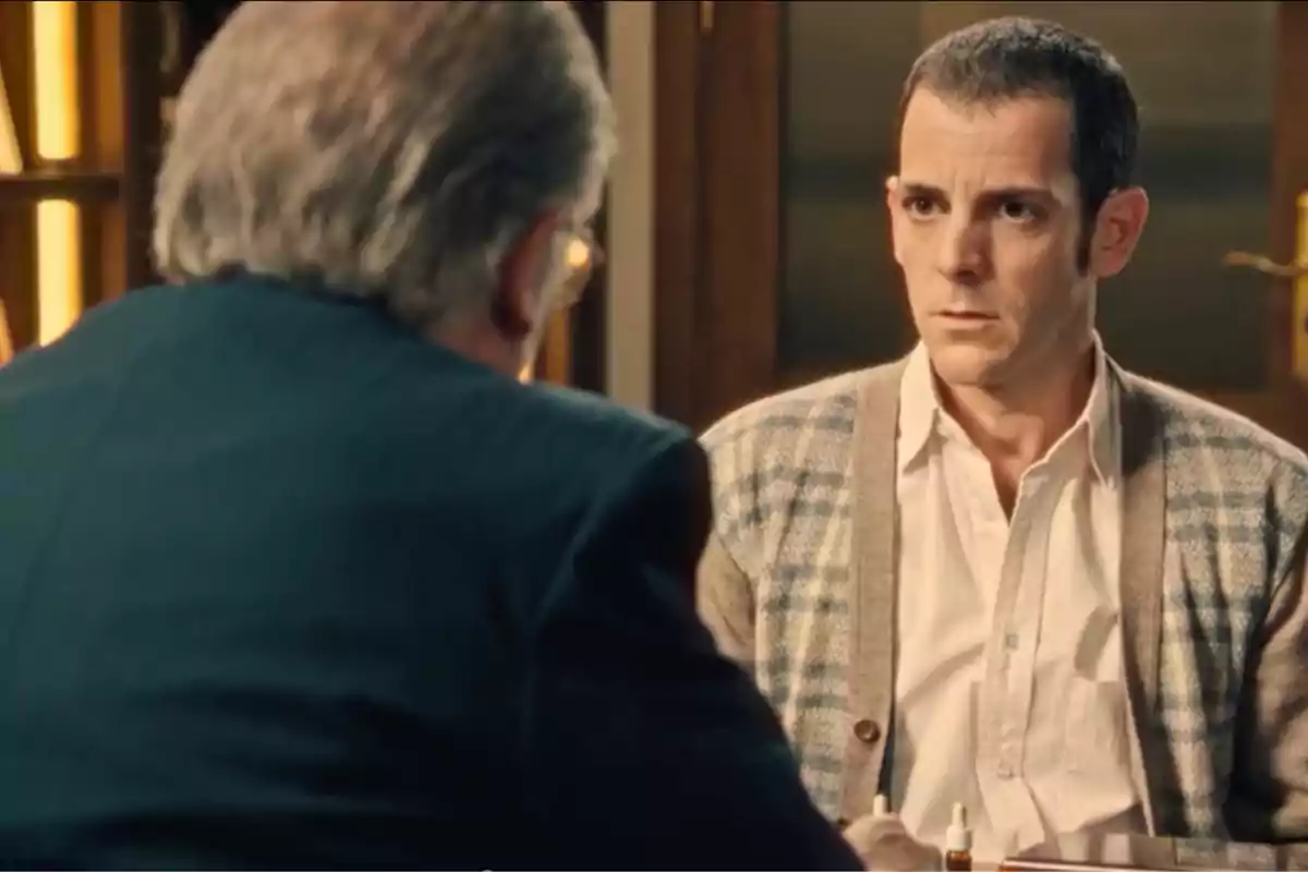 Luis Merino (Guillermo Barrientos) conversa con un hombre sentados frente a frente en una oficina uno de ellos luce serio y lleva un suéter de cuadros Luis Merino (Guillermo Barrientos) conversa con un hombre sentados frente a frente en una oficina uno de ellos luce serio y lleva un suéter de cuadros