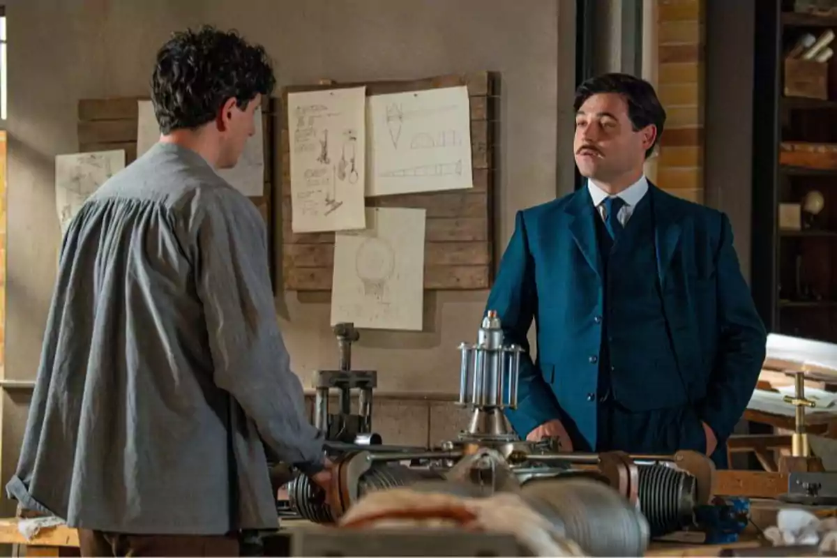 Manuel (Arturo Sancho) conversa con un hombre en un taller con planos y dibujos técnicos en la pared y una máquina antigua sobre la mesa Manuel (Arturo Sancho) conversa con un hombre en un taller con planos y dibujos técnicos en la pared y una máquina antigua sobre la mesa