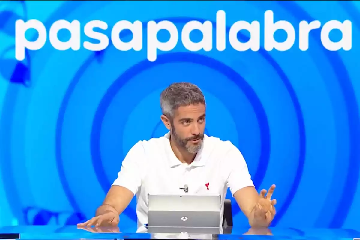 Roberto Leal de cabello canoso y barba sentado frente a una mesa con una tablet, al fondo el logo de Pasapalabra sobre un fondo azul Roberto Leal de cabello canoso y barba sentado frente a una mesa con una tablet, al fondo el logo de Pasapalabra sobre un fondo azul