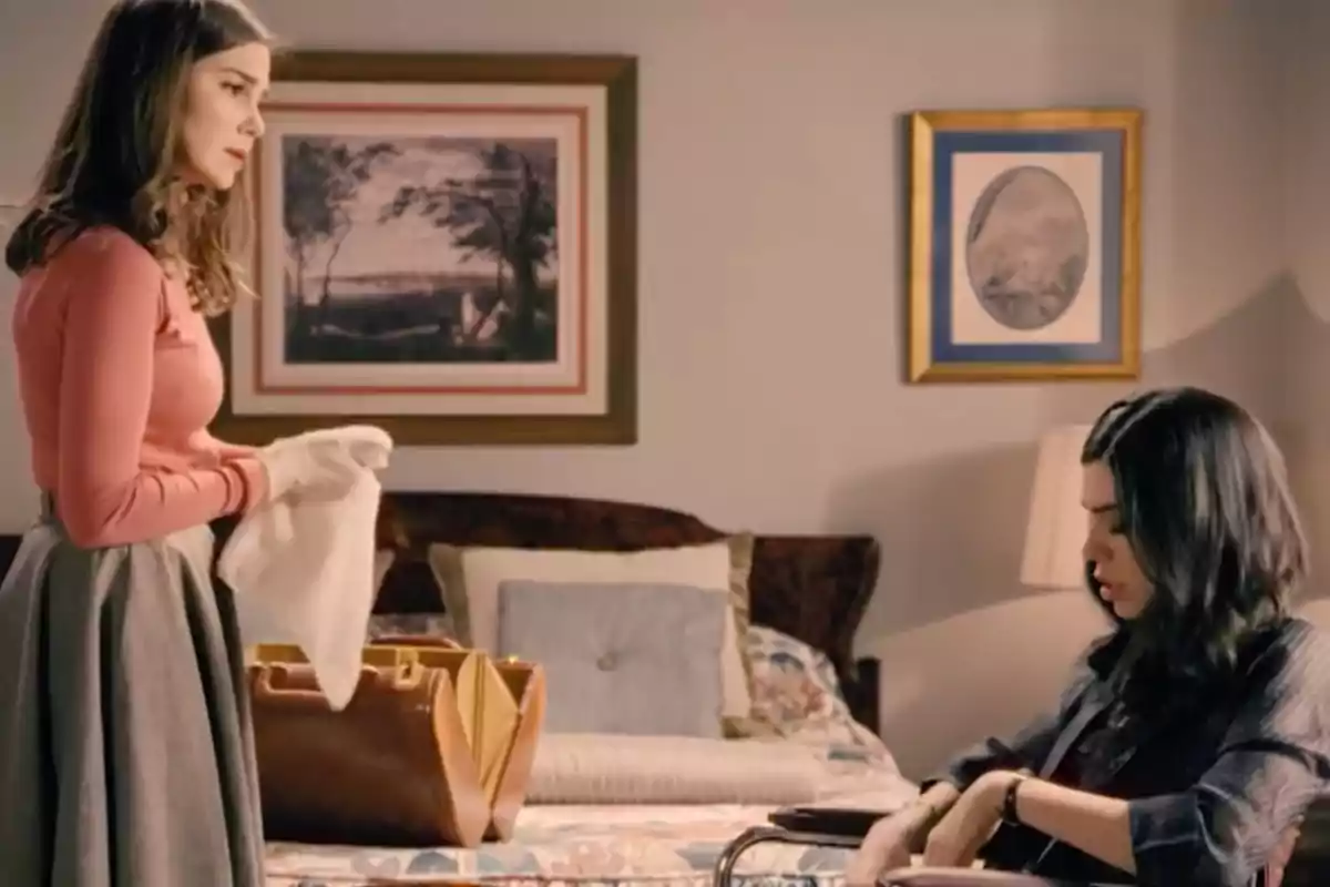 Natalia Sánchez y Roser Tapias están en una habitación, una de pie sosteniendo una prenda blanca y la otra sentada en una silla de ruedas junto a una cama, ambas parecen estar conversando seriamente. Natalia Sánchez y Roser Tapias están en una habitación, una de pie sosteniendo una prenda blanca y la otra sentada en una silla de ruedas junto a una cama, ambas parecen estar conversando seriamente.