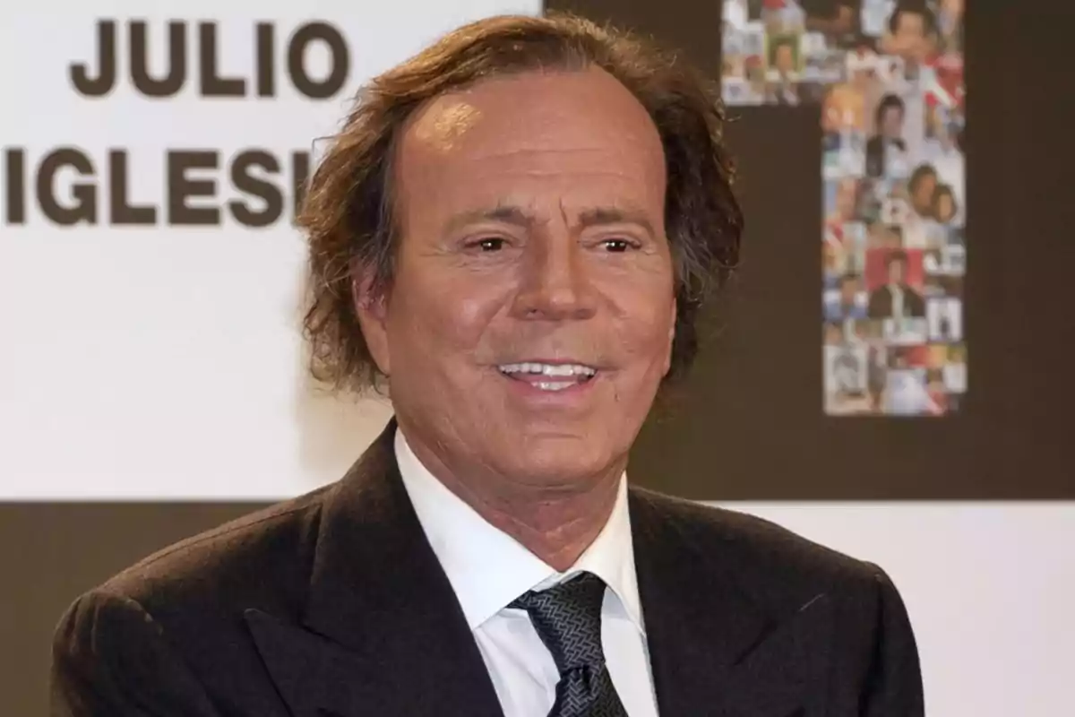 Hombre de cabello castaño y traje oscuro sonriendo frente a un fondo con letras grandes Hombre de cabello castaño y traje oscuro sonriendo frente a un fondo con letras grandes