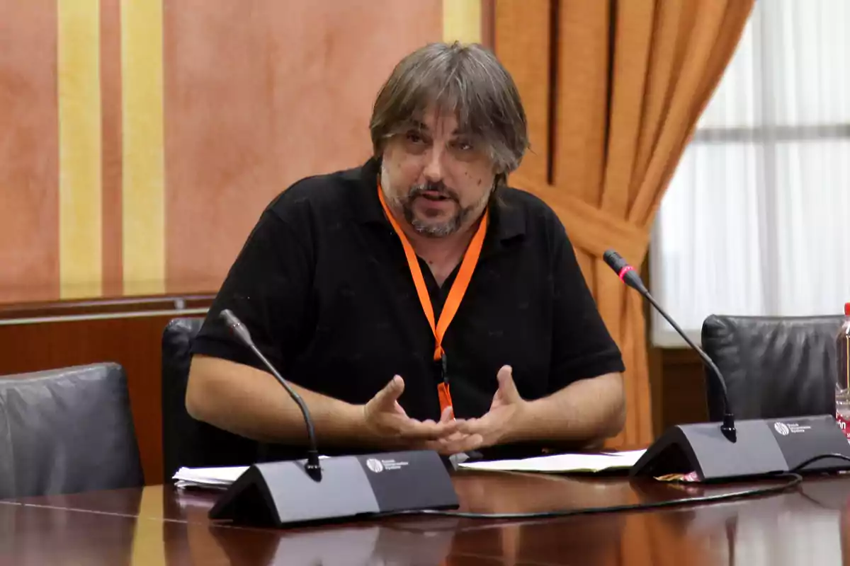 Hombre de cabello canoso y barba sentado en una mesa de reuniones hablando frente a dos micrófonos con cortinas naranjas de fondo Hombre de cabello canoso y barba sentado en una mesa de reuniones hablando frente a dos micrófonos con cortinas naranjas de fondo