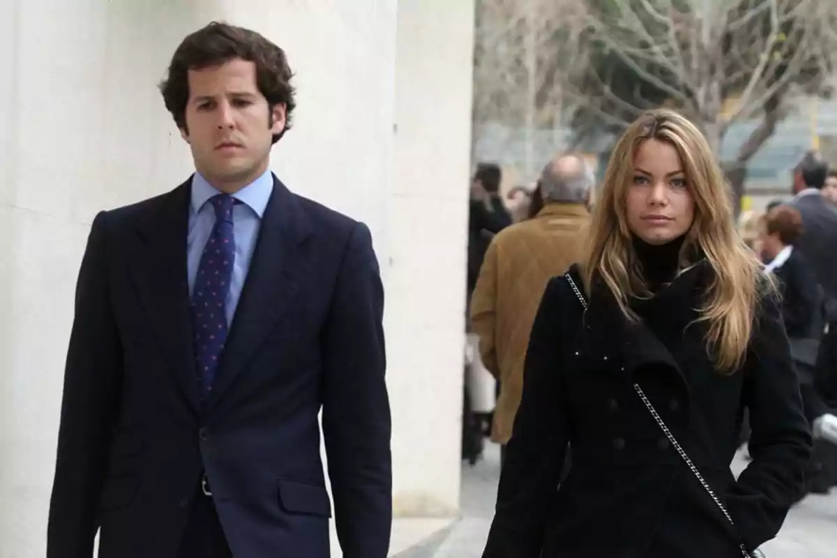 A man in a dark suit and a woman in a black coat walk together down the street in an urban setting with people in the background.