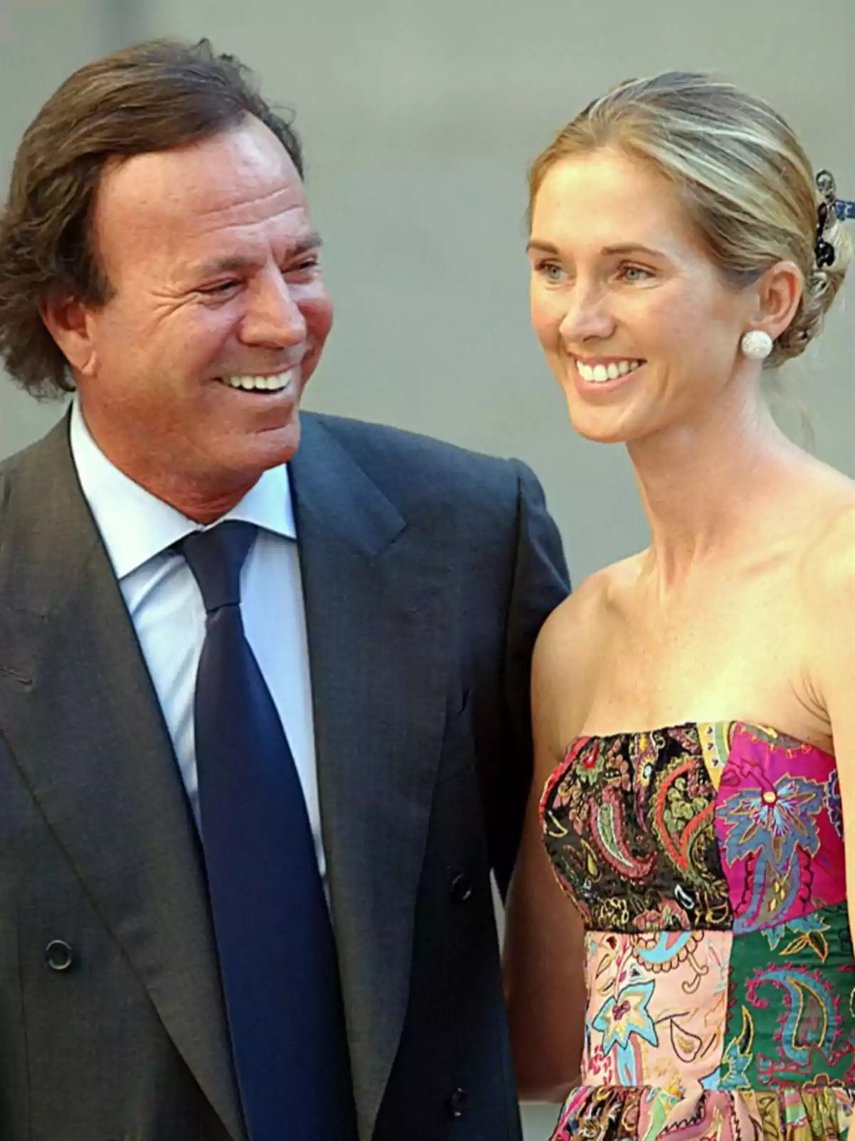 Un hombre de traje oscuro y corbata azul sonríe junto a una mujer rubia con vestido colorido y estampado, ambos posando y mirando hacia el frente Un hombre de traje oscuro y corbata azul sonríe junto a una mujer rubia con vestido colorido y estampado, ambos posando y mirando hacia el frente