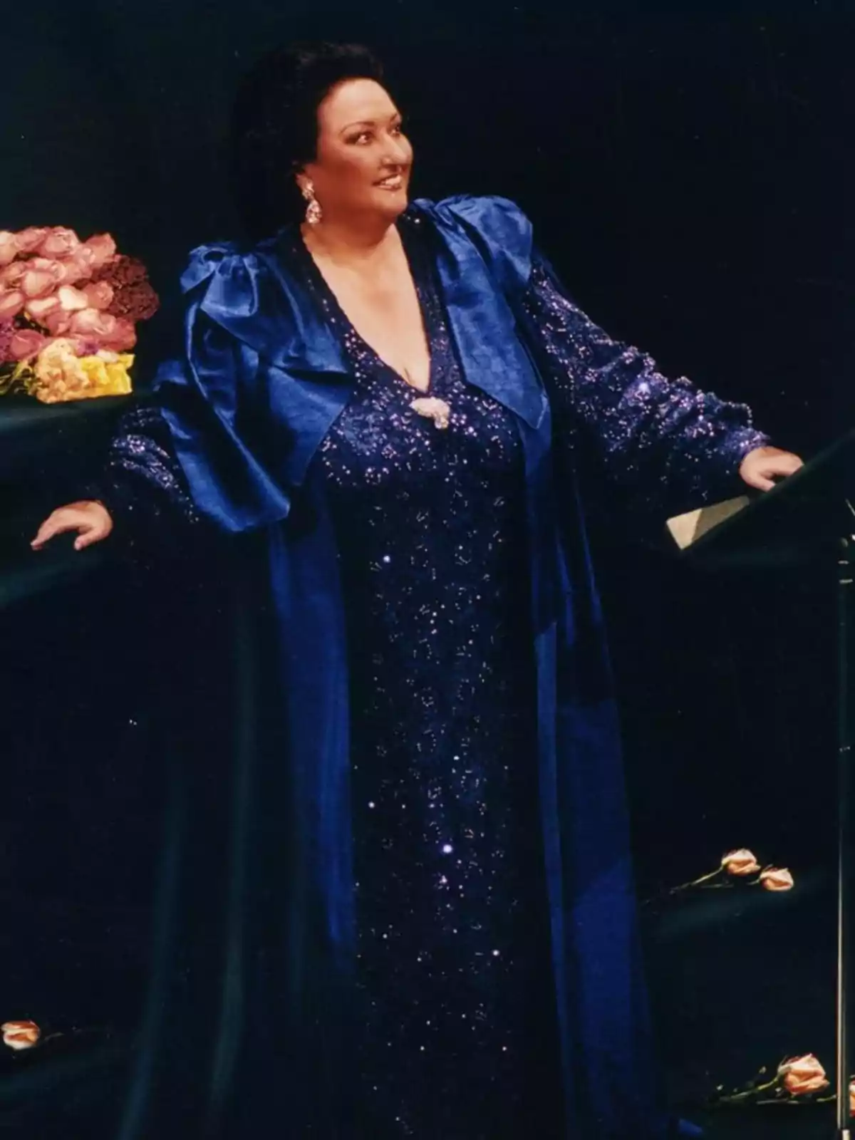 Mujer con vestido azul brillante y capa a juego de pie junto a un ramo de flores rosas sobre una mesa Mujer con vestido azul brillante y capa a juego de pie junto a un ramo de flores rosas sobre una mesa