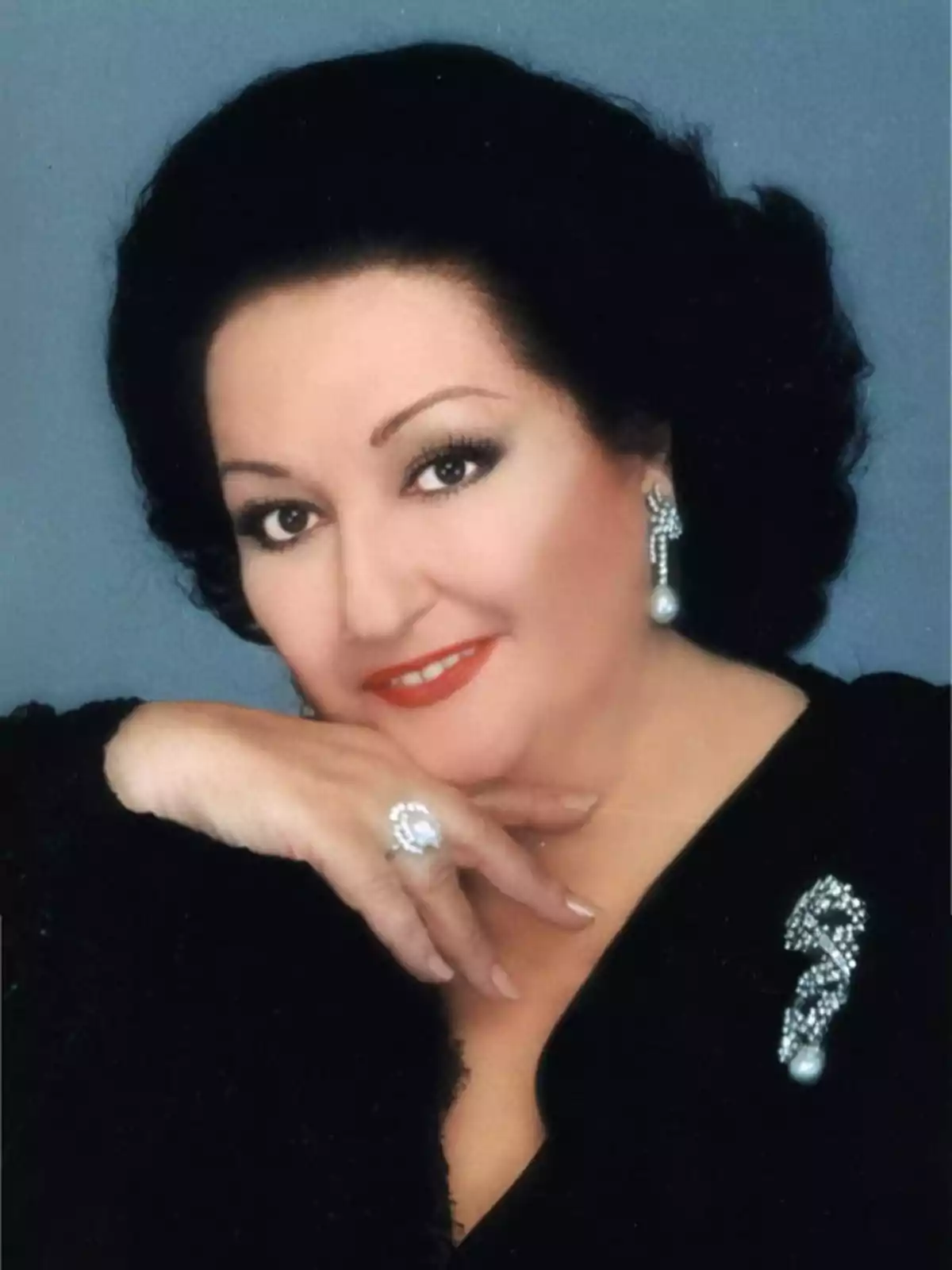 Mujer de cabello oscuro y corto con vestido negro y joyas elegantes, posando con una mano apoyada en el mentón y sonriendo frente a un fondo azul Mujer de cabello oscuro y corto con vestido negro y joyas elegantes, posando con una mano apoyada en el mentón y sonriendo frente a un fondo azul