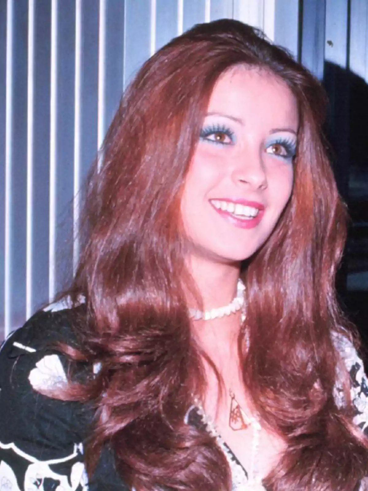 Mujer joven de cabello largo y rojizo sonriendo, con maquillaje llamativo y collar de perlas, frente a un fondo de persianas verticales. Mujer joven de cabello largo y rojizo sonriendo, con maquillaje llamativo y collar de perlas, frente a un fondo de persianas verticales.