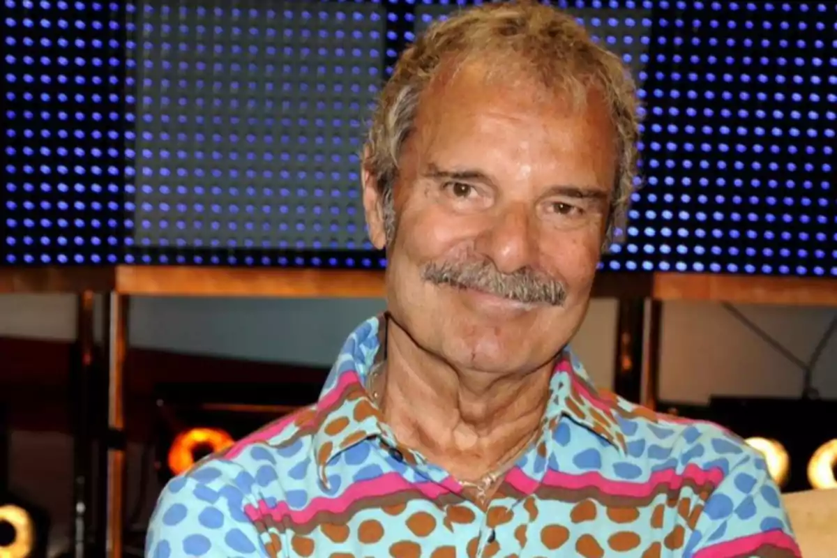 Hombre de cabello canoso y bigote sonríe mientras usa una camisa colorida con estampado de puntos y rayas, fondo con luces azules desenfocadas Hombre de cabello canoso y bigote sonríe mientras usa una camisa colorida con estampado de puntos y rayas, fondo con luces azules desenfocadas