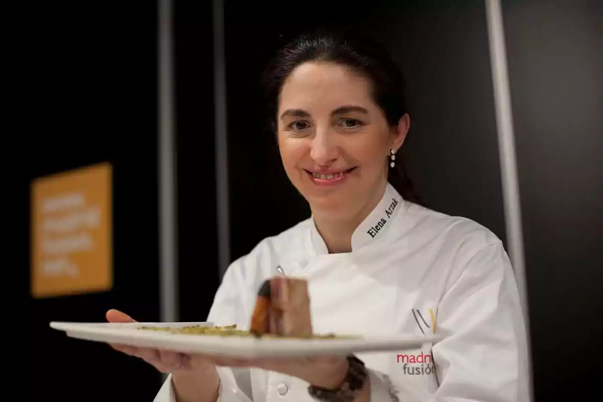 Chef con chaqueta blanca sosteniendo un plato de comida y sonriendo frente a la cámara