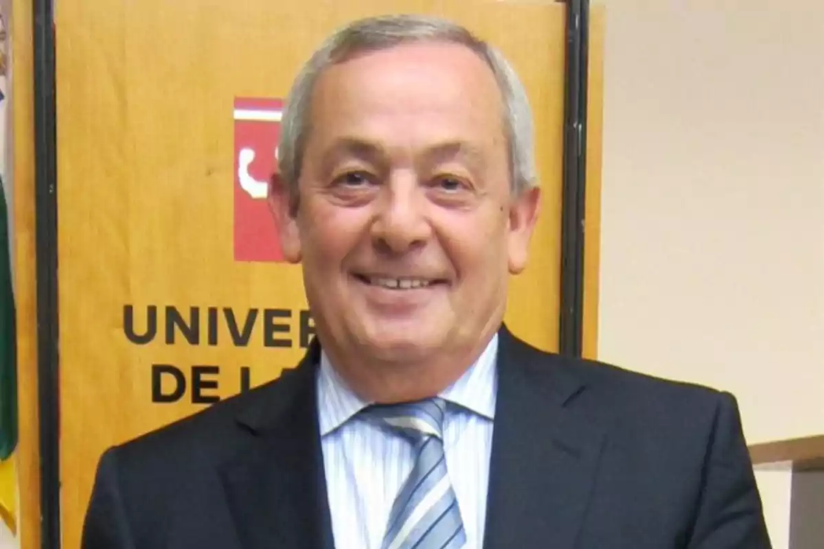 Hombre de cabello canoso y traje oscuro sonriendo frente a un cartel de la universidad Hombre de cabello canoso y traje oscuro sonriendo frente a un cartel de la universidad