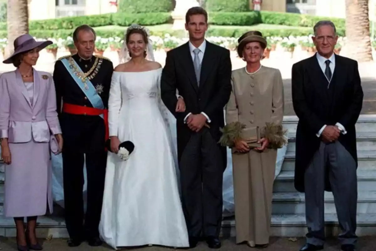 Grupo de seis personas elegantemente vestidas posando para una foto de boda en el exterior, con la novia en el centro usando un vestido blanco y un velo, acompañada por el novio y familiares a ambos lados. Grupo de seis personas elegantemente vestidas posando para una foto de boda en el exterior, con la novia en el centro usando un vestido blanco y un velo, acompañada por el novio y familiares a ambos lados.