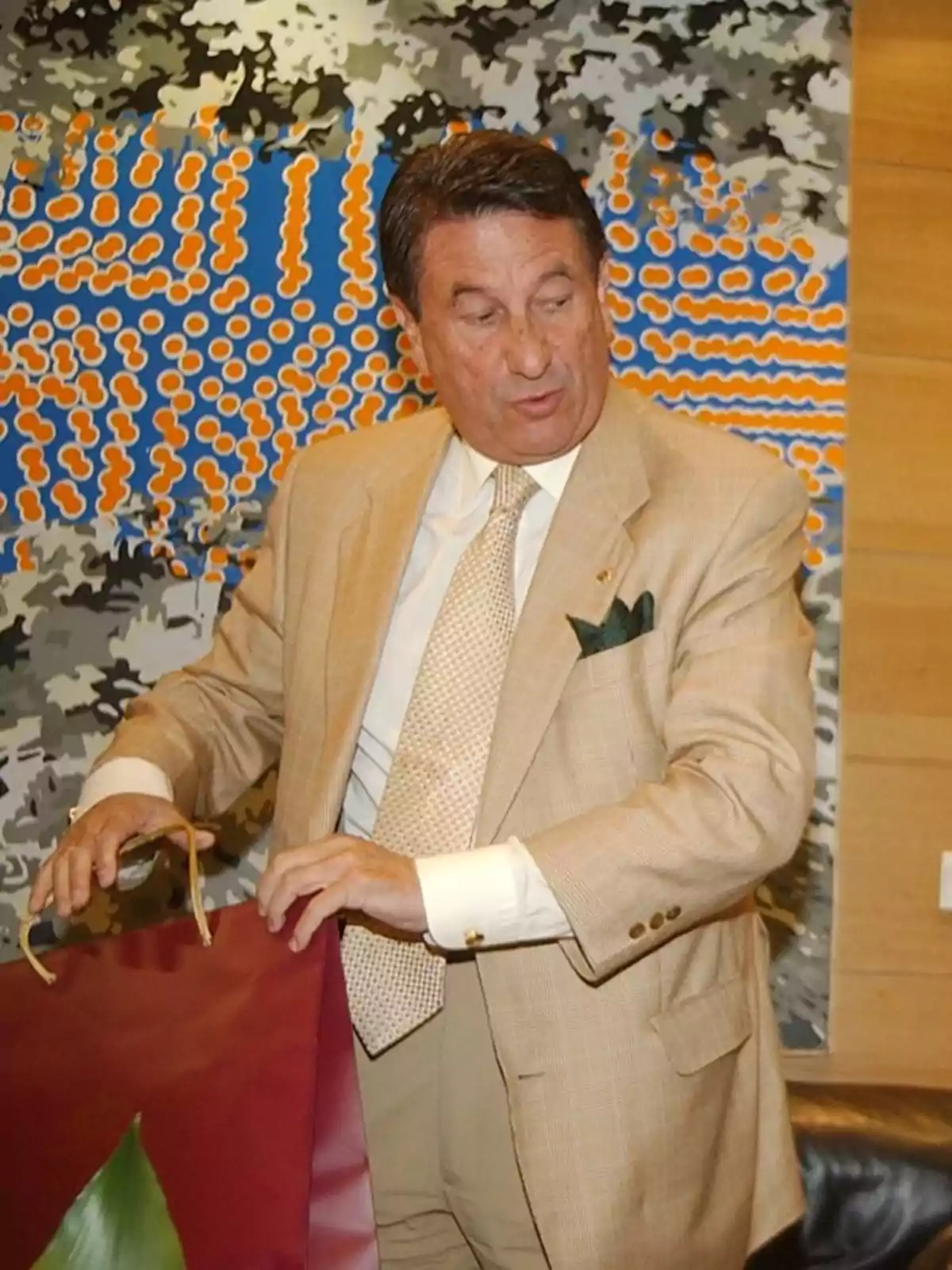 Hombre de cabello oscuro con traje beige y corbata clara sosteniendo una bolsa roja frente a una colorida obra de arte abstracta en la pared Hombre de cabello oscuro con traje beige y corbata clara sosteniendo una bolsa roja frente a una colorida obra de arte abstracta en la pared
