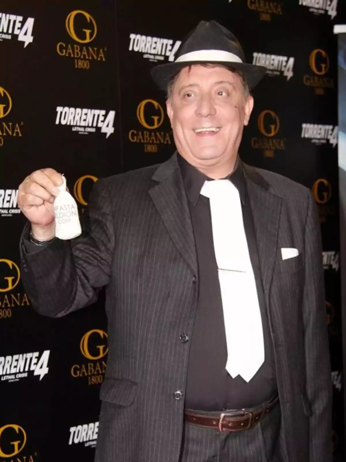 Hombre sonriente con sombrero y corbata blanca posando en un evento con fondo de logotipos de Gabana 1800 y Torrente 4 Hombre sonriente con sombrero y corbata blanca posando en un evento con fondo de logotipos de Gabana 1800 y Torrente 4