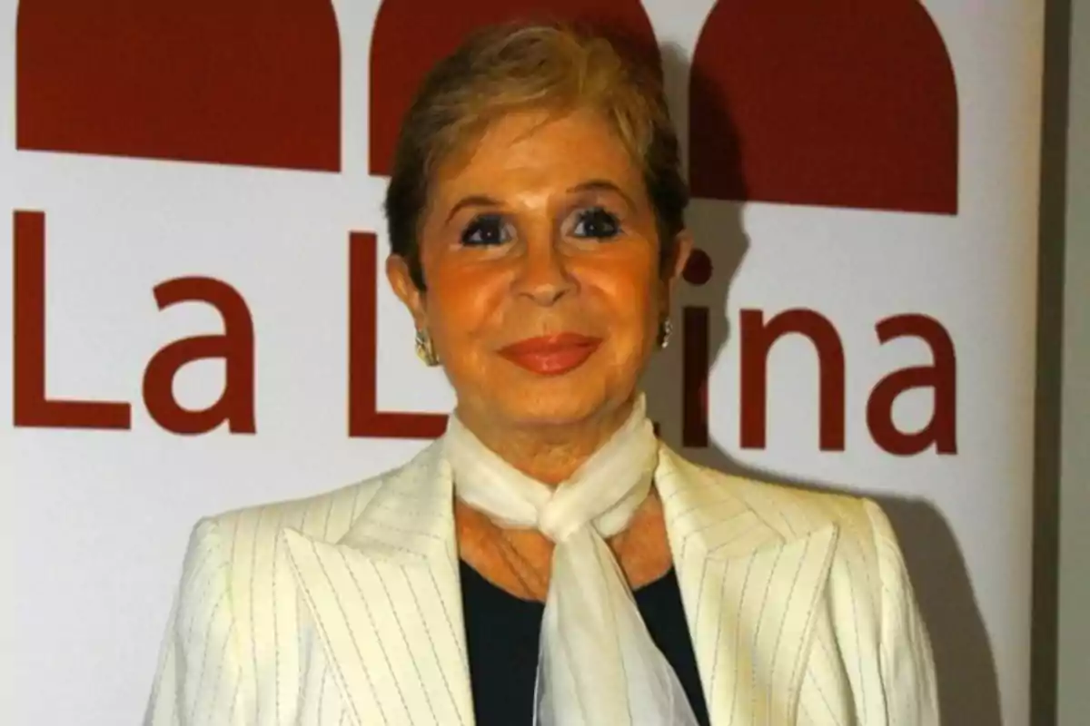 Mujer de cabello corto y rubio vestida con saco claro y bufanda blanca sonriendo frente a un cartel que dice La Lina
