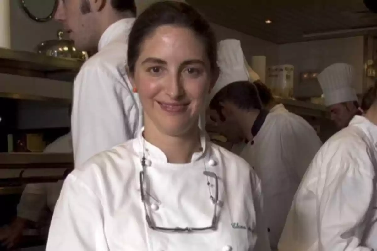 Chef sonriente con bata blanca y gafas colgadas en el cuello en una cocina profesional rodeada de otros cocineros Chef sonriente con bata blanca y gafas colgadas en el cuello en una cocina profesional rodeada de otros cocineros