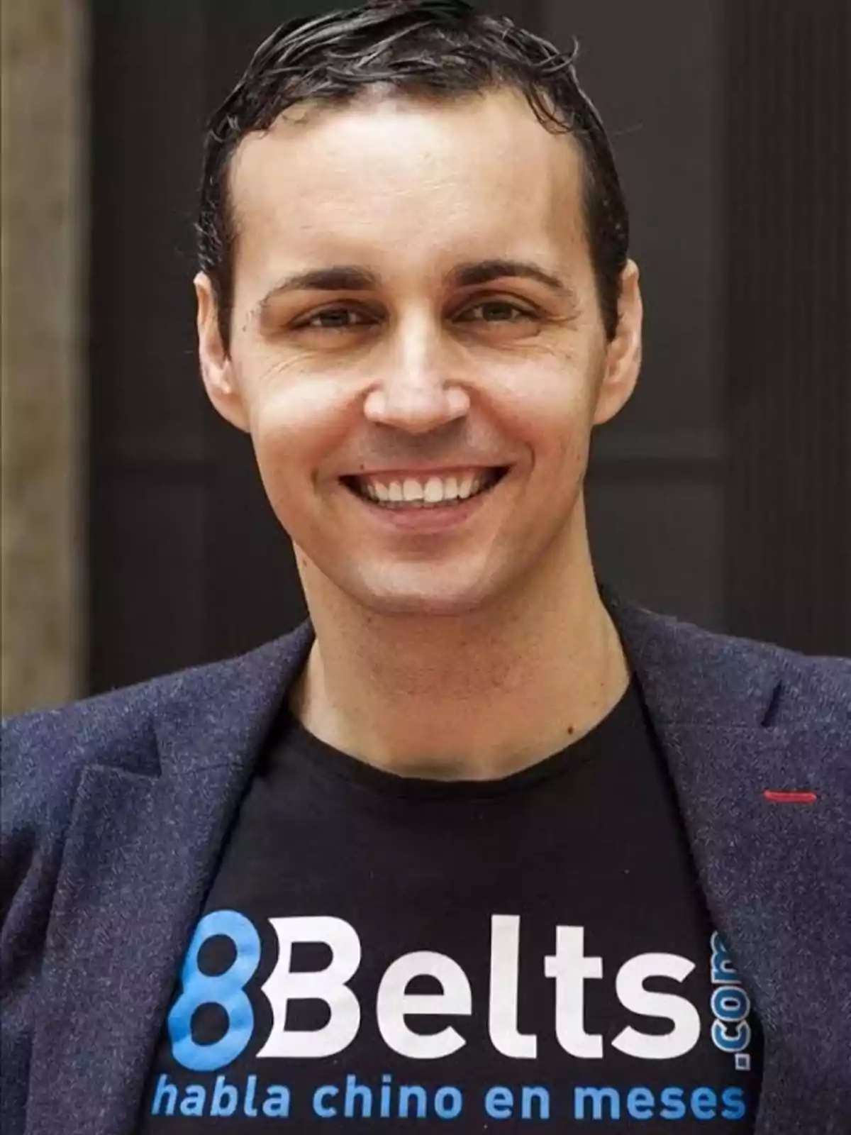 Hombre sonriente con cabello corto y oscuro, vistiendo una camiseta negra con el logo de 8Belts y un saco azul marino Hombre sonriente con cabello corto y oscuro, vistiendo una camiseta negra con el logo de 8Belts y un saco azul marino