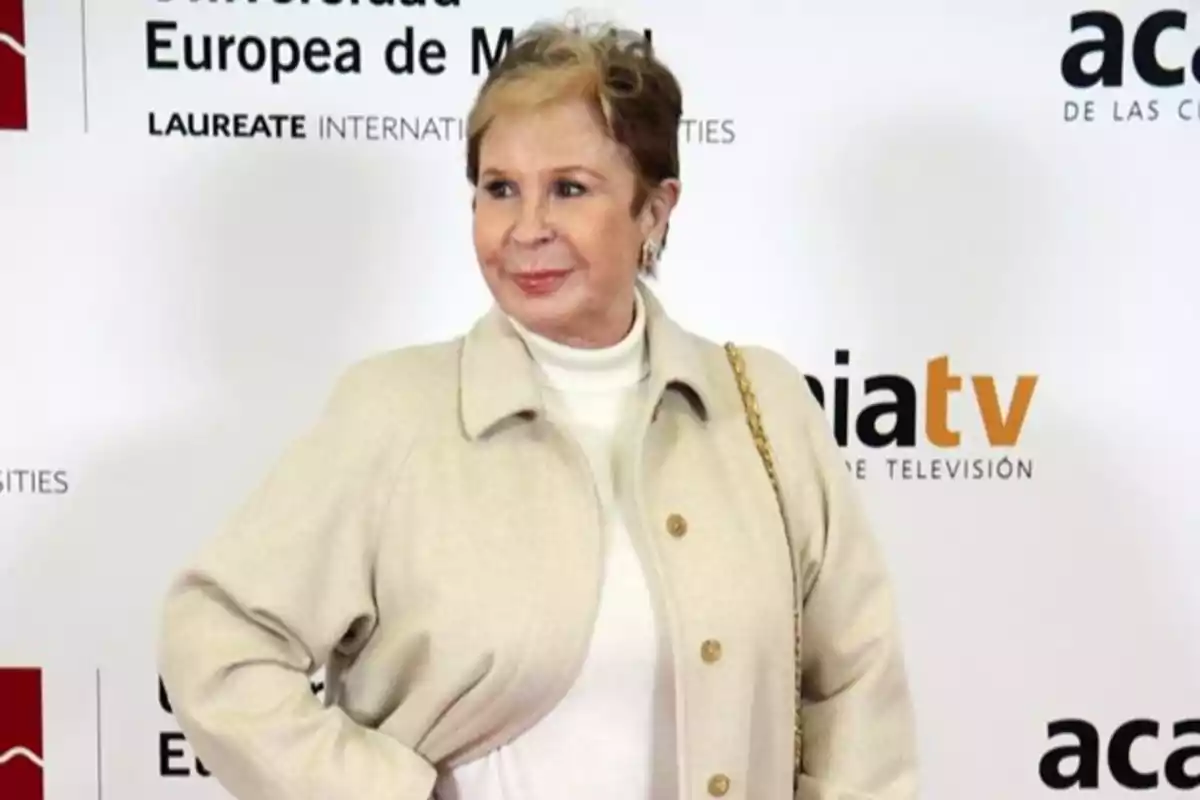 Mujer de cabello corto y rubio vestida con abrigo beige y suéter blanco posando frente a un fondo con logotipos de televisión y universidad