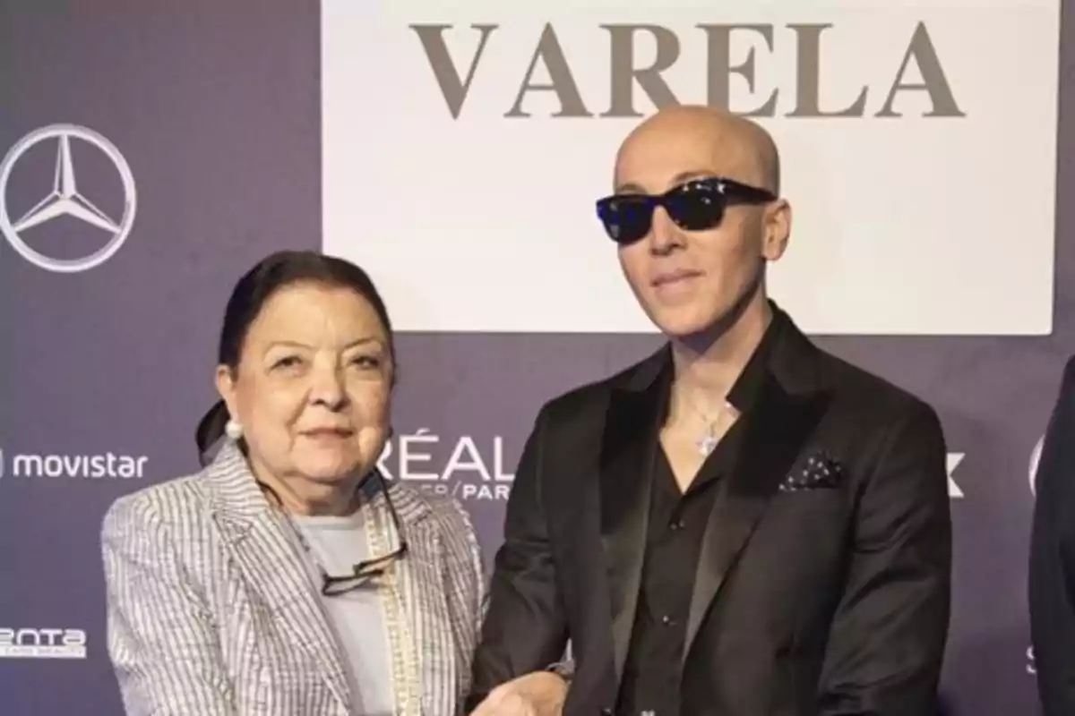 Dos personas posan juntas en un evento formal frente a un fondo con logotipos y la palabra Varela Dos personas posan juntas en un evento formal frente a un fondo con logotipos y la palabra Varela