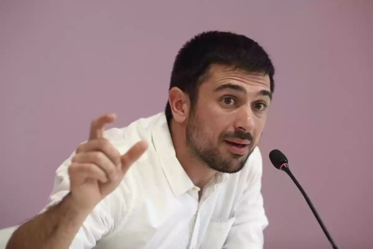 Hombre joven con barba y camisa blanca hablando frente a un micrófono y gesticulando con la mano sobre un fondo lila