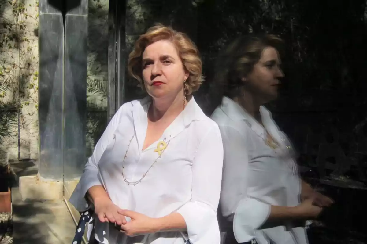 Mujer de cabello corto y rubio vestida con blusa blanca posando junto a una pared de vidrio que refleja su imagen Mujer de cabello corto y rubio vestida con blusa blanca posando junto a una pared de vidrio que refleja su imagen