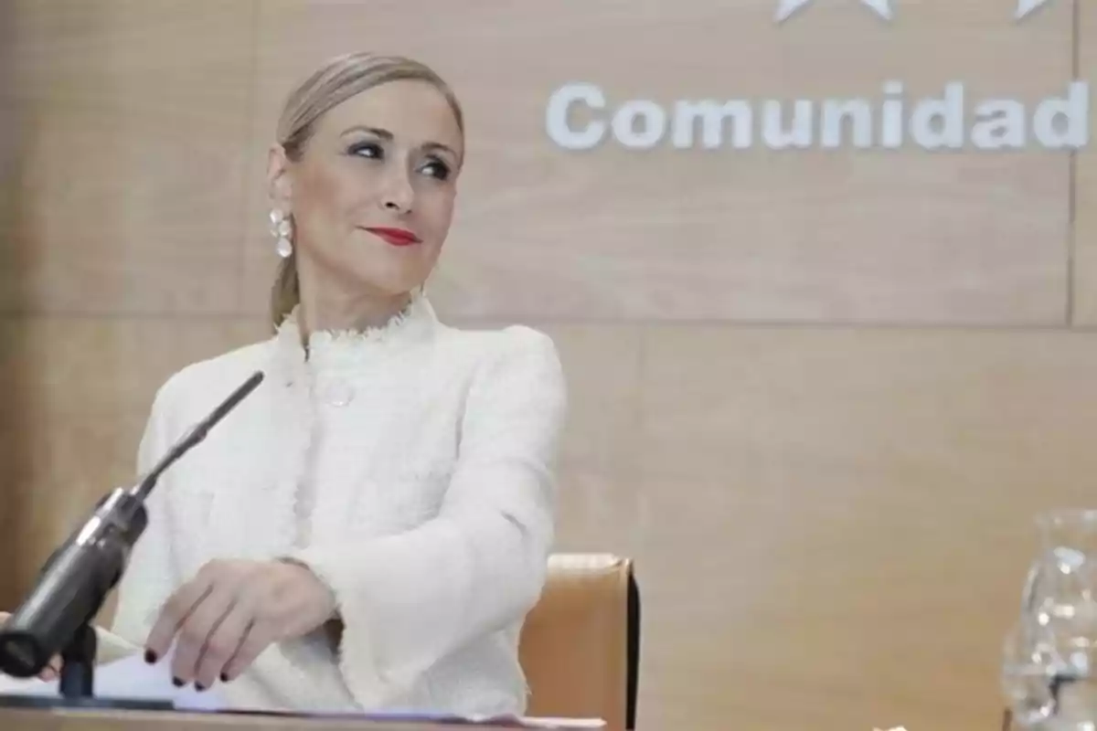 Mujer de cabello rubio recogido y vestida de blanco sentada frente a un micrófono en una sala con fondo de madera y la palabra comunidad escrita detrás Mujer de cabello rubio recogido y vestida de blanco sentada frente a un micrófono en una sala con fondo de madera y la palabra comunidad escrita detrás
