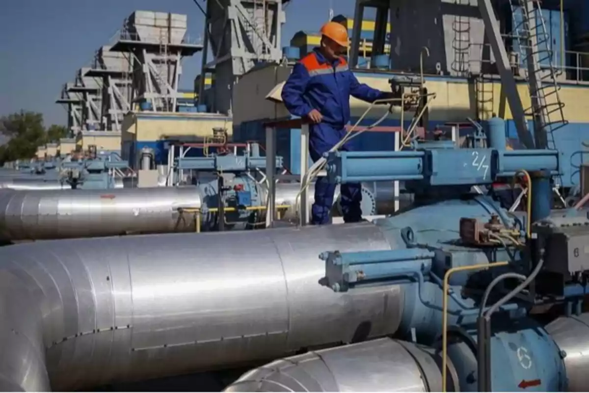 Trabajador con casco y uniforme azul operando maquinaria en una planta industrial con grandes tuberías plateadas y estructuras metálicas al fondo Trabajador con casco y uniforme azul operando maquinaria en una planta industrial con grandes tuberías plateadas y estructuras metálicas al fondo