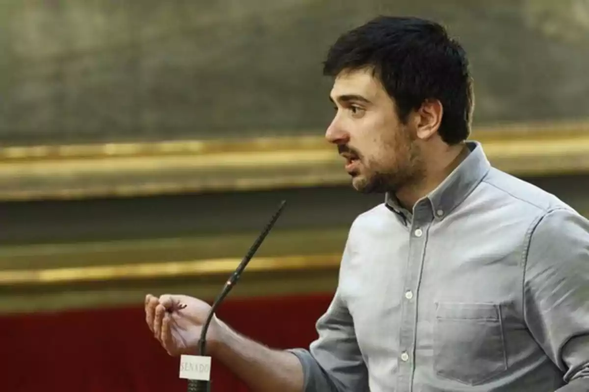 Hombre joven de cabello oscuro y barba corta hablando frente a un micrófono en un recinto formal Hombre joven de cabello oscuro y barba corta hablando frente a un micrófono en un recinto formal