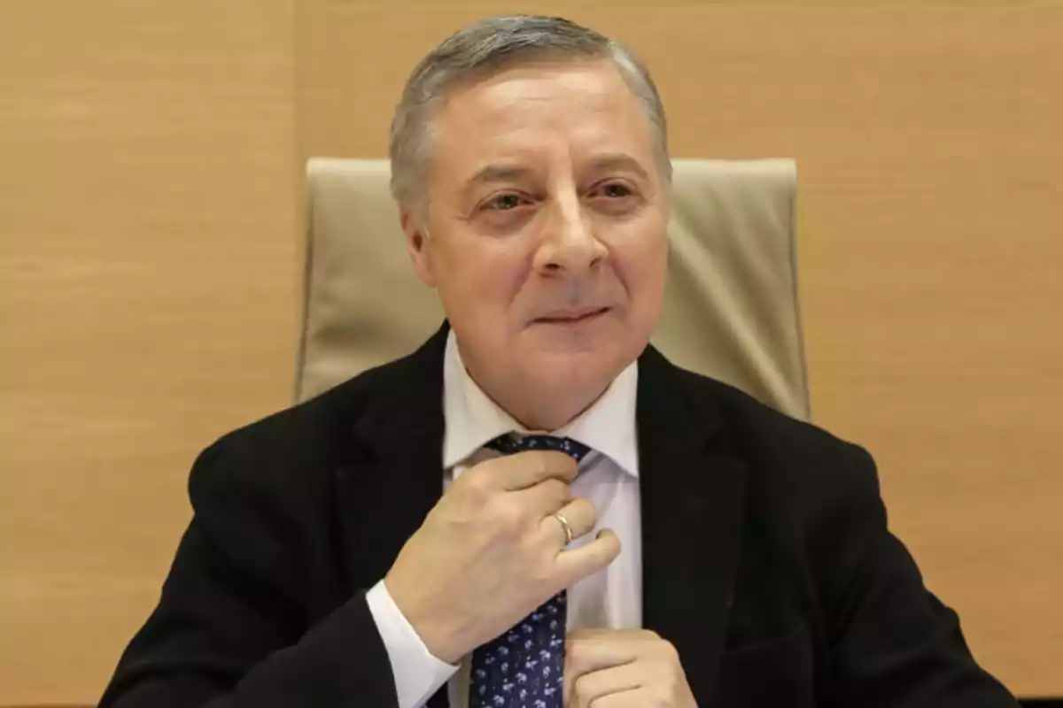 Hombre de cabello canoso sentado en una silla ajustándose la corbata con una expresión seria y fondo de madera clara Hombre de cabello canoso sentado en una silla ajustándose la corbata con una expresión seria y fondo de madera clara