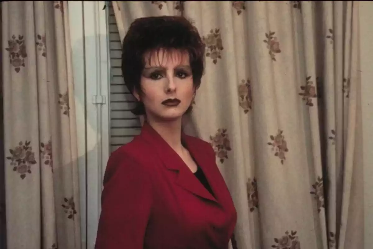 Mujer con maquillaje oscuro y peinado voluminoso usando un saco rojo frente a cortinas beige con flores. Mujer con maquillaje oscuro y peinado voluminoso usando un saco rojo frente a cortinas beige con flores.