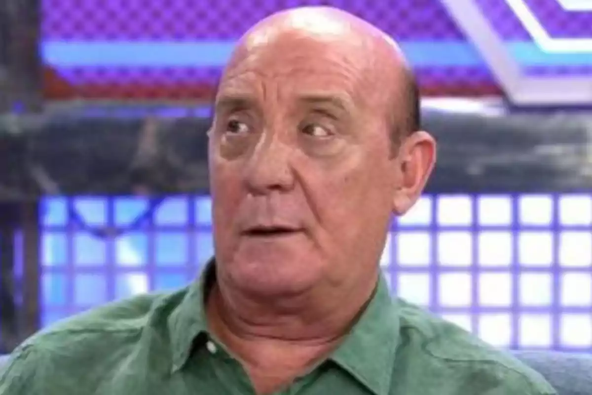 Hombre calvo con camisa verde mirando hacia un lado en un set de televisión con fondo azul y morado. Hombre calvo con camisa verde mirando hacia un lado en un set de televisión con fondo azul y morado.
