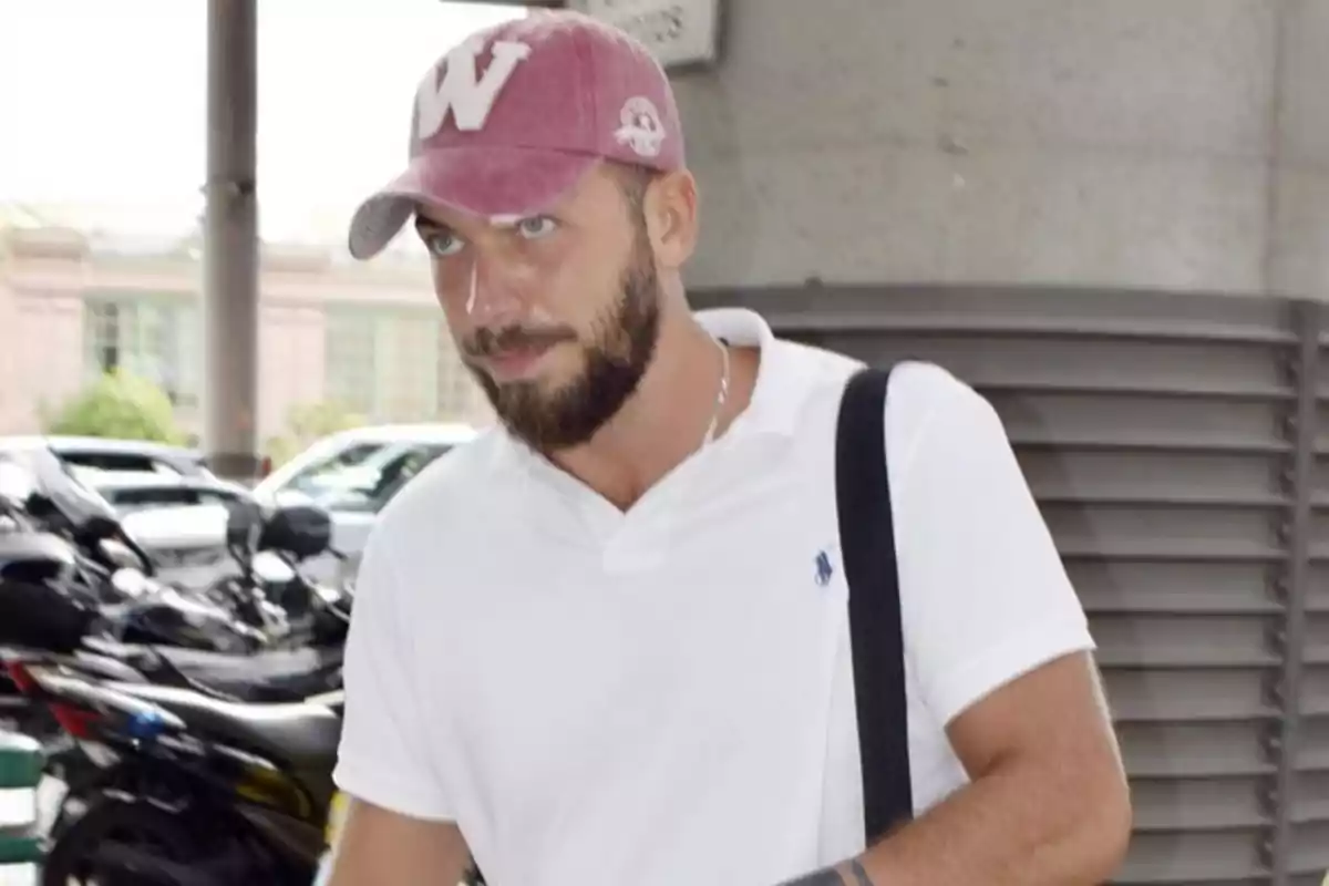 Hombre con barba y gorra rosa caminando al aire libre con una camiseta polo blanca y una mochila negra Hombre con barba y gorra rosa caminando al aire libre con una camiseta polo blanca y una mochila negra