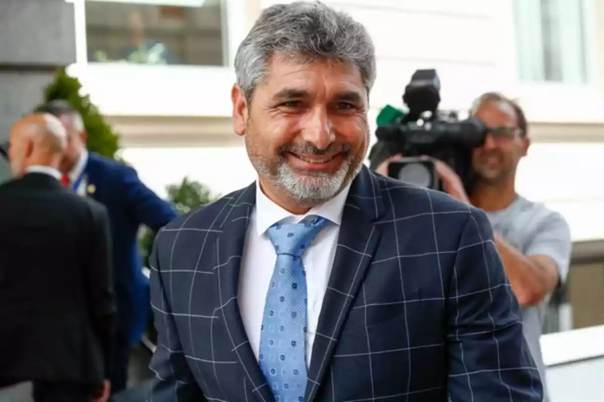 Hombre de cabello canoso y barba sonríe mientras viste un traje azul con cuadros y corbata celeste en un entorno urbano con personas y una cámara de video al fondo