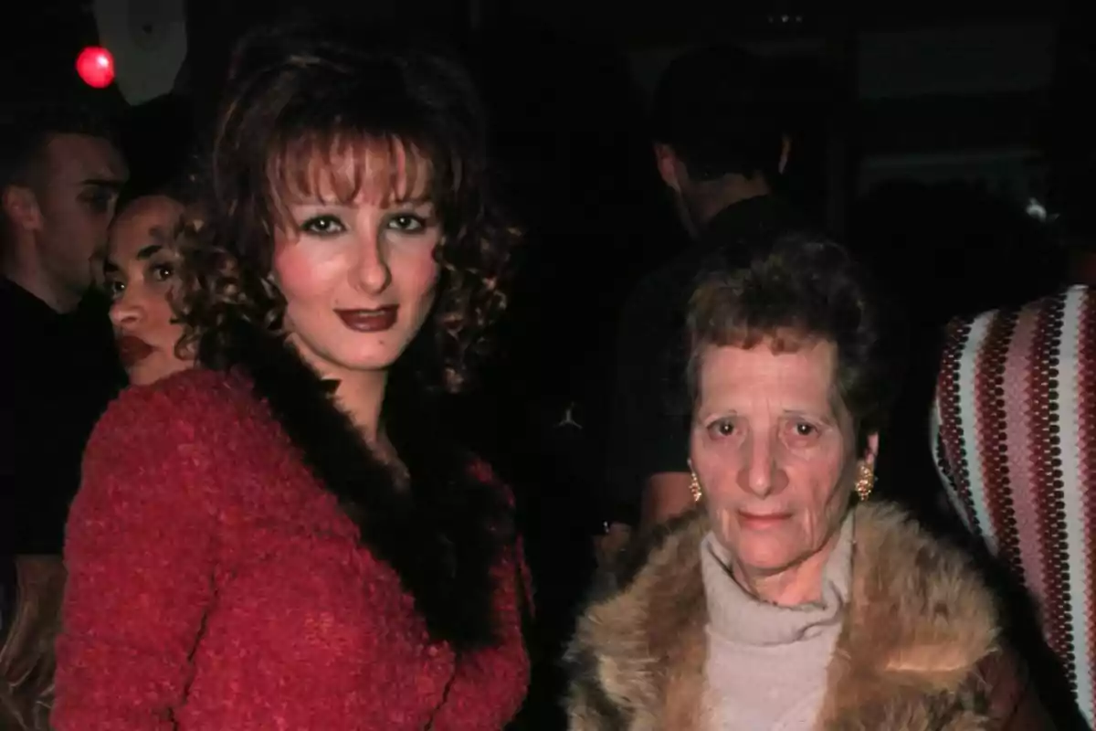 Dos mujeres posan juntas en un lugar oscuro con varias personas al fondo, ambas llevan ropa de invierno elegante Dos mujeres posan juntas en un lugar oscuro con varias personas al fondo, ambas llevan ropa de invierno elegante