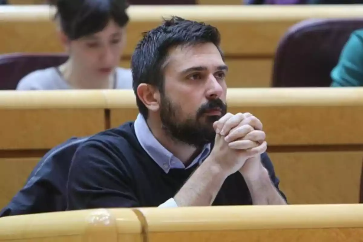 Hombre con barba y cabello corto sentado en una sala de conferencias con las manos entrelazadas y expresión pensativa Hombre con barba y cabello corto sentado en una sala de conferencias con las manos entrelazadas y expresión pensativa