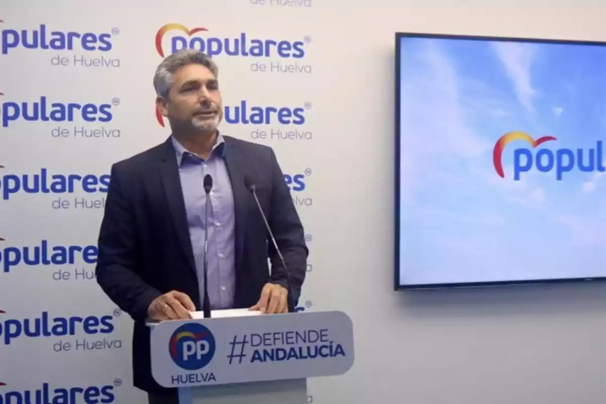 Un hombre de traje habla en una rueda de prensa del Partido Popular de Huelva con un fondo y una pantalla que muestran el logo del partido y el lema defiende Andalucía Un hombre de traje habla en una rueda de prensa del Partido Popular de Huelva con un fondo y una pantalla que muestran el logo del partido y el lema defiende Andalucía