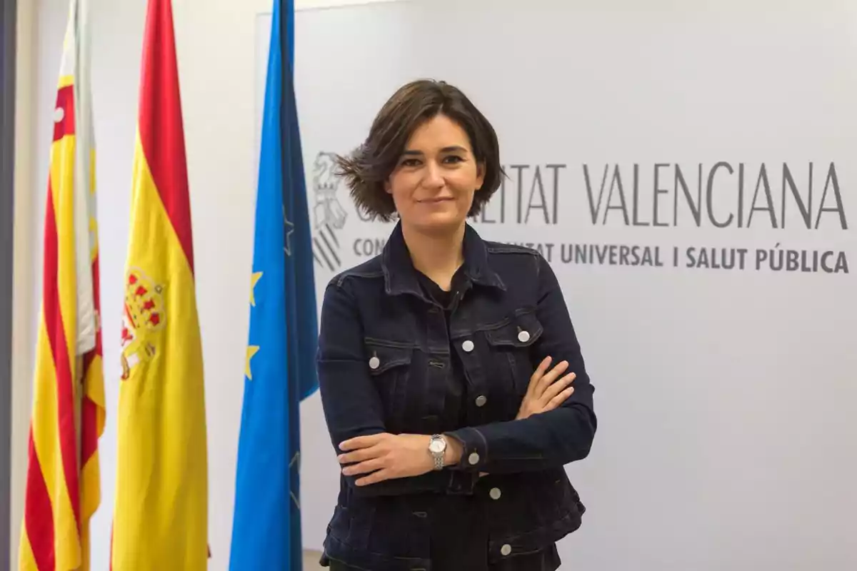 Mujer de cabello corto y oscuro con chaqueta de mezclilla posando con los brazos cruzados frente a un fondo institucional con banderas y un cartel que dice Generalitat Valenciana Mujer de cabello corto y oscuro con chaqueta de mezclilla posando con los brazos cruzados frente a un fondo institucional con banderas y un cartel que dice Generalitat Valenciana