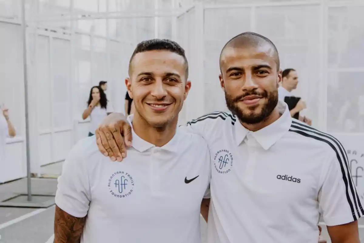 Dos hombres sonríen y posan juntos usando camisetas blancas de la misma fundación en un ambiente al aire libre Dos hombres sonríen y posan juntos usando camisetas blancas de la misma fundación en un ambiente al aire libre