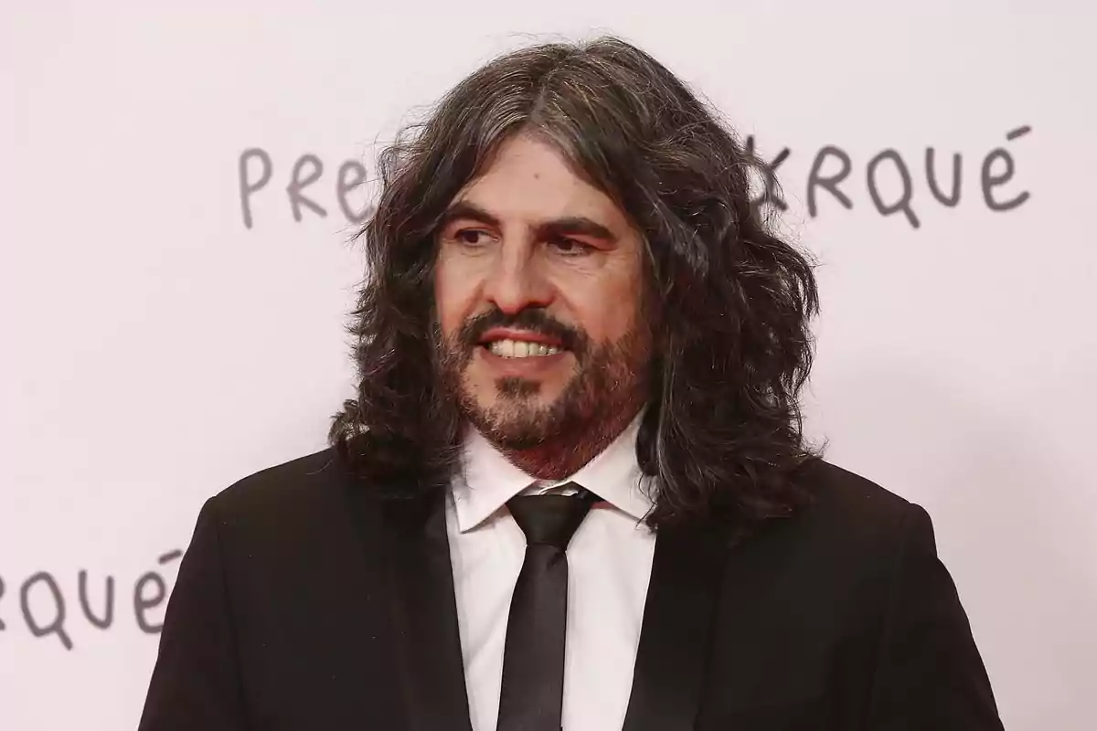 Hombre de cabello largo y barba con traje negro y corbata posando frente a un fondo claro con letras desenfocadas Hombre de cabello largo y barba con traje negro y corbata posando frente a un fondo claro con letras desenfocadas