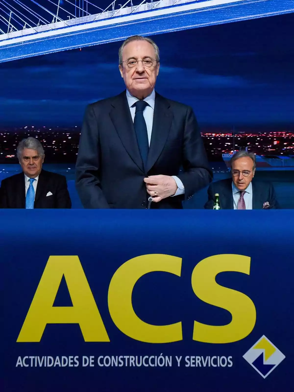 Tres hombres con traje en una conferencia de la empresa ACS Actividades de Construcción y Servicios con un puente iluminado de fondo Tres hombres con traje en una conferencia de la empresa ACS Actividades de Construcción y Servicios con un puente iluminado de fondo