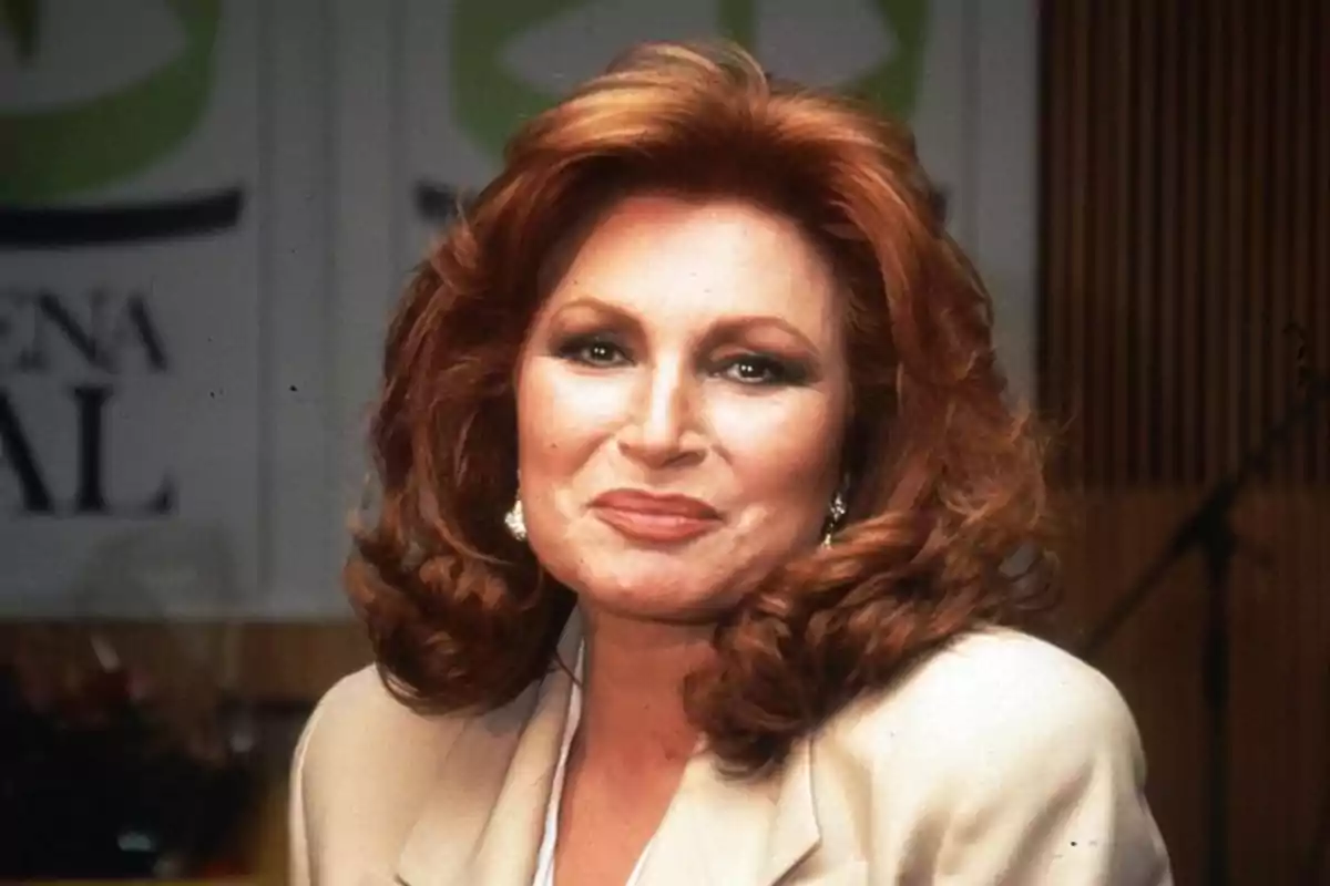 Mujer de cabello pelirrojo y ondulado con maquillaje elegante y vestimenta formal sonriendo frente a un fondo desenfocado
